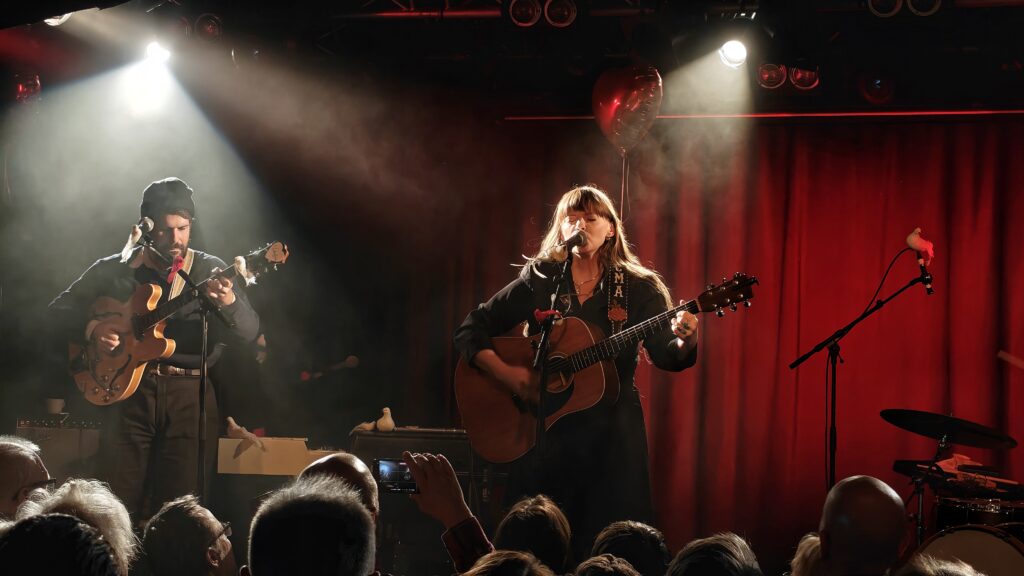 En kvinna med gitarr sjunger i spotlightljus med rätt draperi i bakgrunden, till vänster om henne en medmusiker.
