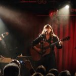 En kvinna med gitarr sjunger i spotlightljus med rätt draperi i bakgrunden, till vänster om henne en medmusiker.