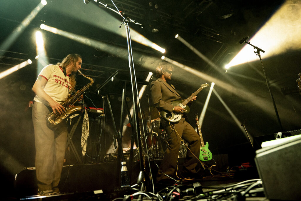 En saxofonist och en man i solgalsögon med elgitarr på en scen.