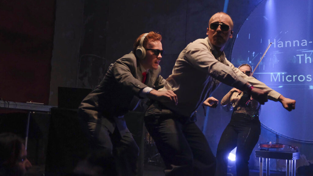 Två män gör rodd-rörelser med en kvinnlig violinist i bakgrunden på scenen