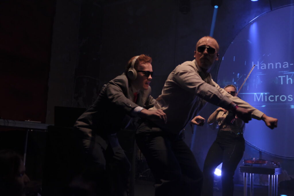 Två män gör rodd-rörelser med en kvinnlig violinist i bakgrunden på scenen.