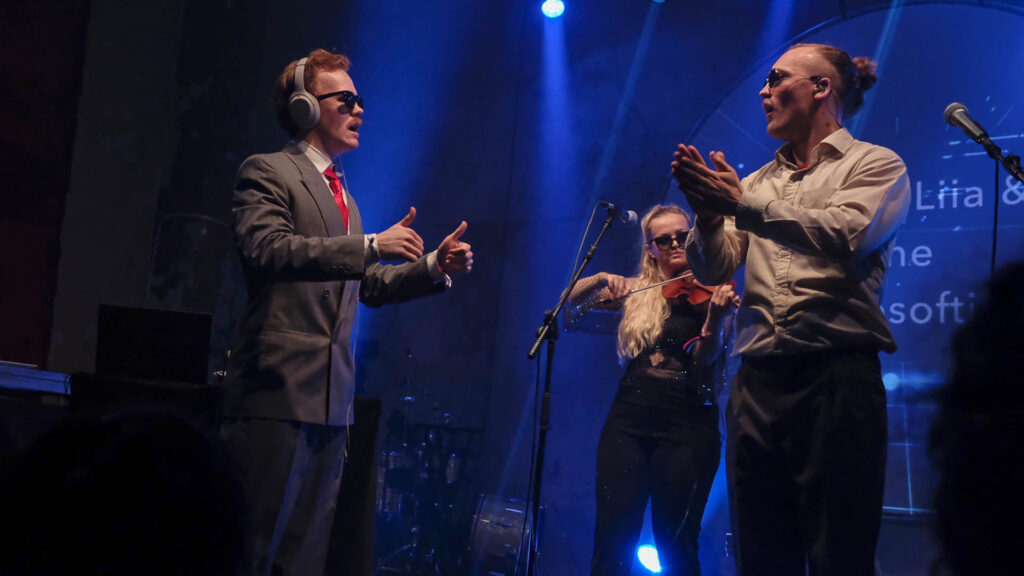 Två unga män med solglasögon tittar mot varandra på scenen, den ena gör tummarna upp. I bakgrunden en kvinna med blont hår som spelar fiol.